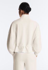Personne aux cheveux foncés en chignon portant une veste ample beige clair à col montant et un pantalon assorti, debout, mains dans les poches, vue de dos.