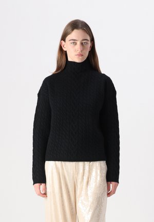 Pull en tricot noir avec un col montant, présentant un motif en cable et une coupe décontractée, associé à un pantalon large en matériau léger et brillant.