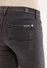 Primer plano de un bolsillo trasero de jeans de mezclilla gris oscuro con etiquetas de tela de marca y un diseño bordado en la cintura de un maniquí.