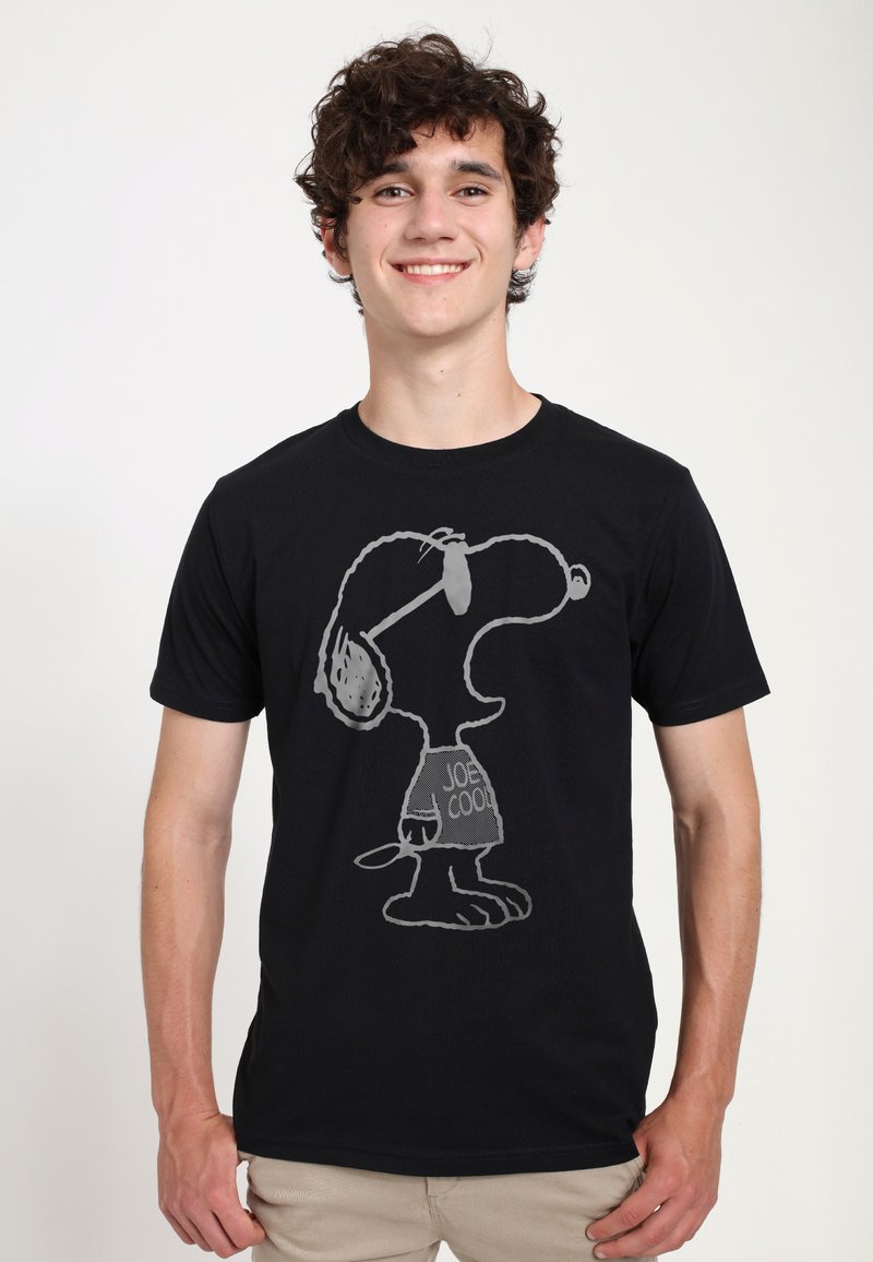 Jeune homme souriant, portant un t-shirt noir avec un personnage de dessin animé Snoopy gris et le texte « Joe Cool ».