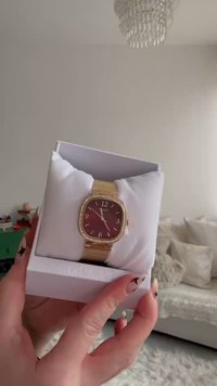 Ρολόι σε χρυσή απόχρωση με τετράγωνο μπορντό καντράν, χρυστό πλέγμα μπράτσου και διακοσμητικά με κρύσταλλα. Παρουσιάζεται πάνω σε λευκό μαξιλαράκι μέσα σε κουτί.