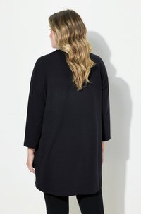 Tunika nera a maniche lunghe con una vestibilità morbida, caratterizzata da un orlo posteriore arrotondato e una texture liscia. Indossata con leggings neri.