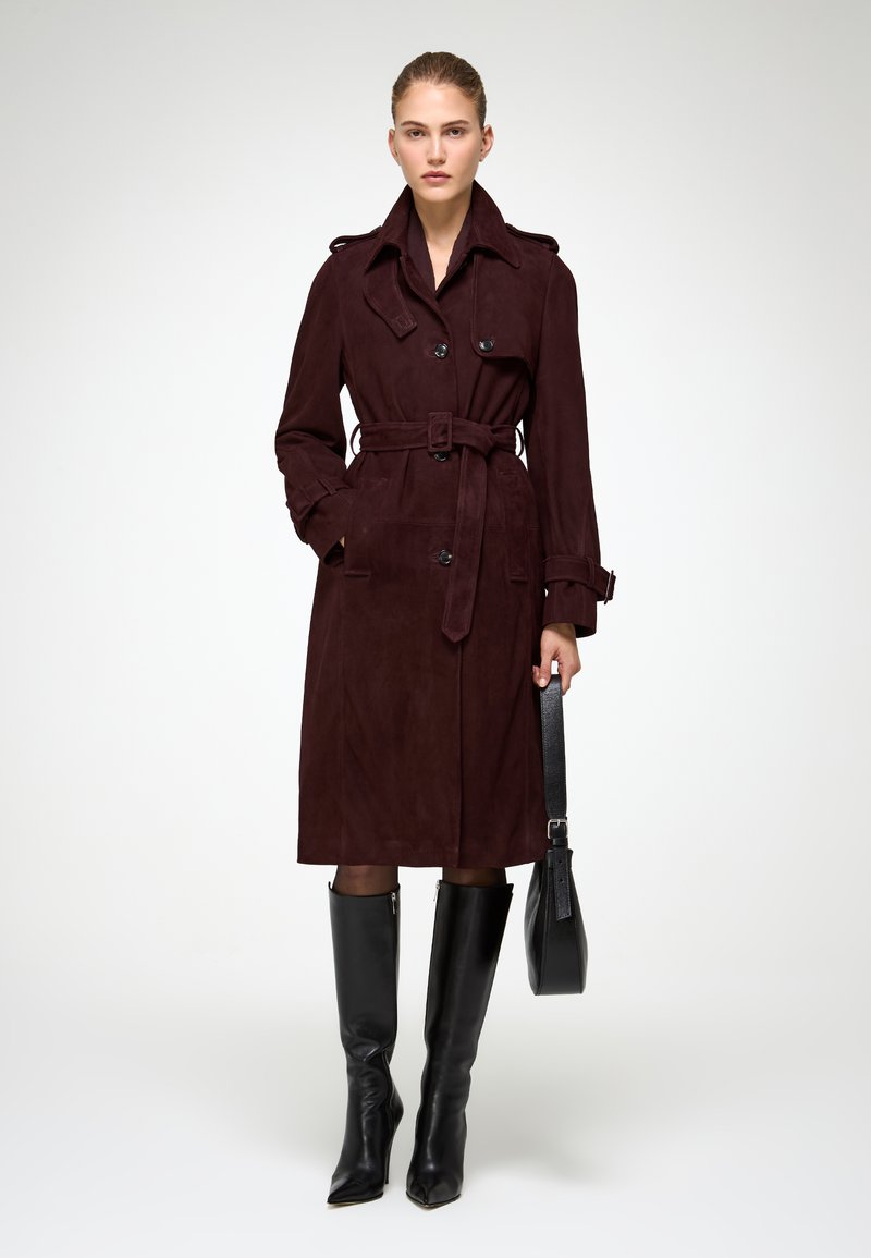 Burgundský semišový trench coat s opaskom v páse, dvojradovými gombíkmi a bočnými vreckami. Model má na sebe čierne čižmy po kolená a drží čiernu tašku.