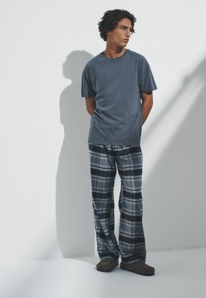 Junger Mann mit lockigem Haar, der ein dunkelblaues T-Shirt, graue und schwarze karierte Hosen und braune Schuhe trägt und vor einem weißen Hintergrund steht.