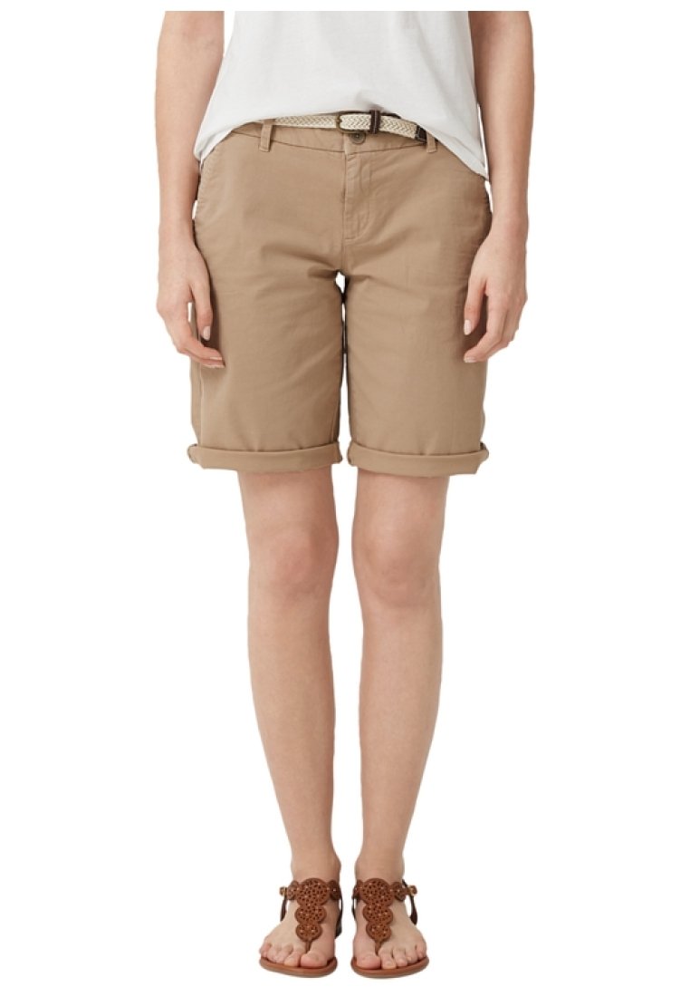 Femme portant un short beige jusqu'aux genoux, retroussé, avec une ceinture blanche, un haut blanc et des sandales décoratives marron, se tenant devant un fond blanc.