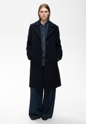 Frau steht in einem schwarzen, geknöpften Mantel über einem Jeanshemd und weiten blauen Jeanshosen mit schwarzen Schuhen, Hände in den Manteltaschen.