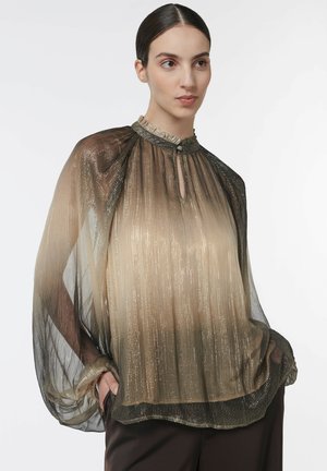 Femme aux cheveux foncés rassemblés en chignon bas, vêtue d’un chemisier ombré métallisé transparent à manches bouffantes et d’un pantalon foncé, debout devant un fond uni.