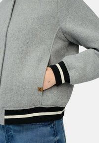 Graue Wollmischungjacke mit einer Knopfleiste, gerippten schwarz-weißen Bündchen und einer seitlichen Tasche. Besitzt ein kleines braunes Etikett an der Seite.
