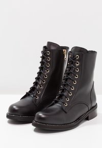 Černé kožené šněrovací boty s kulatou špičkou, zlatou kovovou aplikací, bočním zipem a texturovaným švem podél podešve. Obsahují polstrovanou vnitřní podšívku.