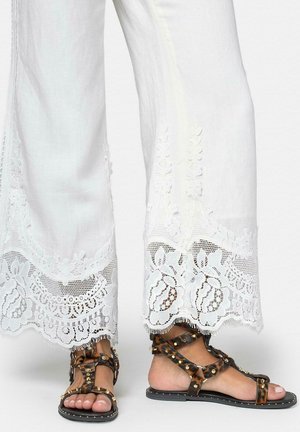 Pantalones blancos de pierna ancha con ribete de encaje floral, combinados con sandalias planas marrones con tachuelas, sobre un fondo liso.