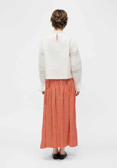 Άτομο με κοντά σγουρά μαλλιά φοράει λευκό πουκάμισο με μακριά μανίκια και δαντελένιες λεπτομέρειες και μακριά κόκκινη και λευκή καρό φούστα, γυρισμένο από πίσω.