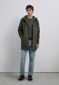 En ung man som bär en olivgrön regnjacka med huva, grått tröja, ljusblåjeans och mörkbruna skor, stående mot en enkel bakgrund.