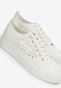 Weiße Sneakers mit Spitzenmuster und niedriger Form, weißen Schnürsenkeln und Gummisohlen, schräg nach rechts auf einem weißen Hintergrund.