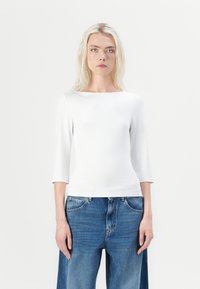 Hvit topp med trekvart ermer, tettsittende design, glatt tekstur. Kombinert med løse blå denimjeans, med knapp og lommer.
