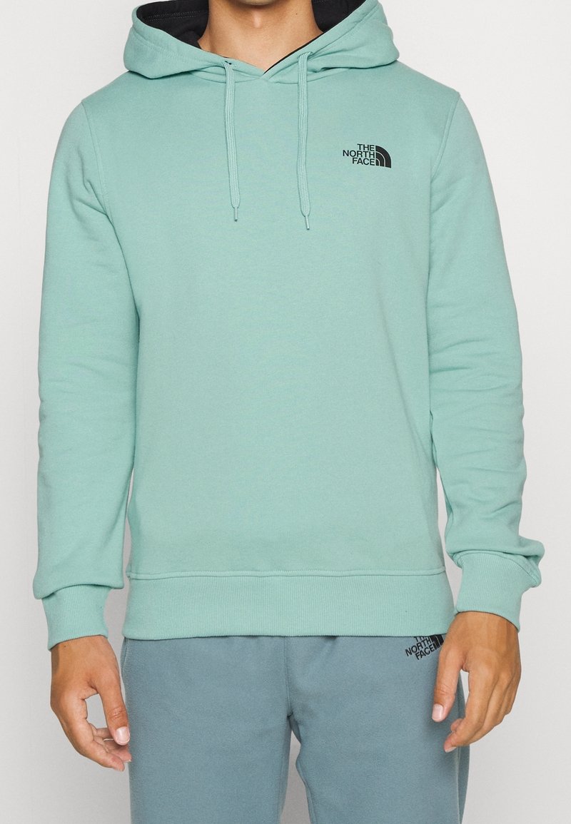Personne portant un sweat à capuche vert menthe The North Face et un pantalon de survêtement assorti, debout les bras détendus le long du corps.