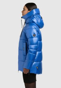 Blå puffjacka med hög krage, fickor med dragkedja och texturerad vaddering. Tillverkad i ett glänsande syntetmaterial med en slimmad design.