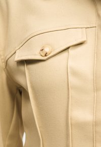 Τσέπη από μπεζ υφασμάτινο σακάκι με καλύπτρα ασφαλισμένη με ένα μοναδικό μαρμάρινο μπεζ και καφέ κουμπί, λεπτομερώς ραμμένη με ακριβή ραφή.