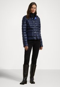 Μπλε σκούρο μπουφάν τύπου puffer με οριζόντια σχεδίαση καπιτονέ, κουμπιά με σούστα και δύο μπροστινές τσέπες. Φορεμένο με μαύρο εφαρμοστό παντελόνι και μαύρες μπότες μέχρι το γόνατο.