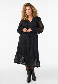 Čierne čipkované šaty s dlhými nadýchanými rukávmi, výstrihom do V a zaväzovaním v páse. Majú vrstvený lem a gombíky na prednej strane.