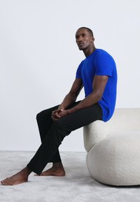 T-shirt blu in cotone, abbinato a pantaloni neri. Soggetto seduto su un divano bianco rotondo e testurizzato, a piedi nudi su un tappeto grigio chiaro.