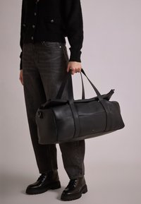 Musta nahkainen duffel-laukku, jossa on kaksoiskahvat, minimalistiinen muotoilu, sileä pinta ja hienovarainen brändäys, jota kantaa henkilö tummissa vaatteissa.