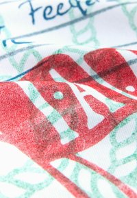 Gros plan sur un tissu avec une illustration rouge de dinde, un motif de feuilles bleu sarcelle, et un texte bleu partiel sur fond blanc.