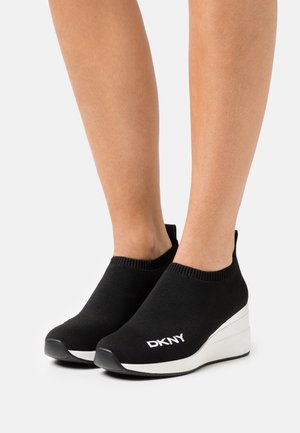 Sorte strikkede slip-on sko med en hvid kile-sål. Har en ribbet ankel og "DKNY" logo i hvid på siden. Glat tekstur.