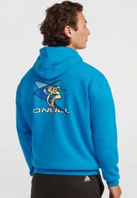 Homme portant un sweat à capuche O'Neill bleu vif avec un grand graphisme stylisé de surfeur dans le dos, associé à un pantalon noir sur fond uni.