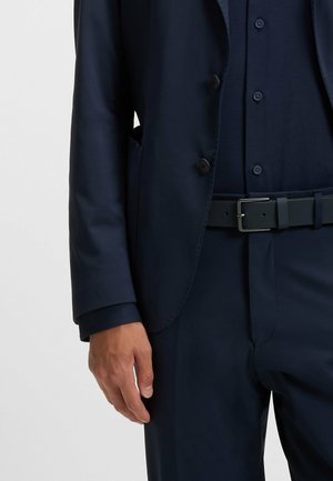 Giacca da abito blu navy in tessuto liscio, con due bottoni, rever a punta e tasche laterali, abbinata a una camicia e una cintura coordinate.