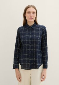 Chemise à carreaux bleu marine avec des coutures blanches, manches longues, col et ourlet arrondi. Tissu léger avec une texture lisse.