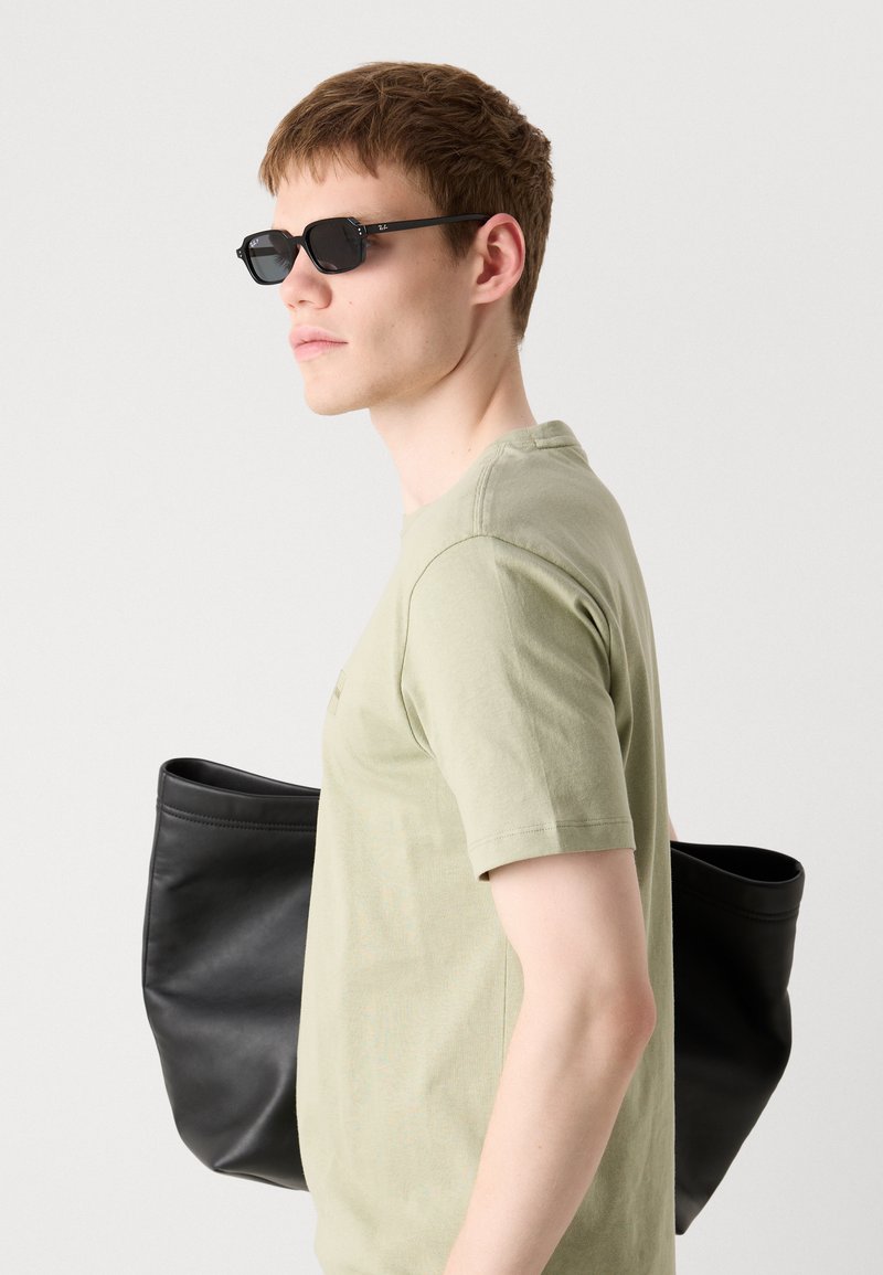 Des lunettes de soleil noires, un t-shirt en coton vert olive clair et un sac fourre-tout en cuir noir sont présentés. La silhouette est moderne et minimaliste.
