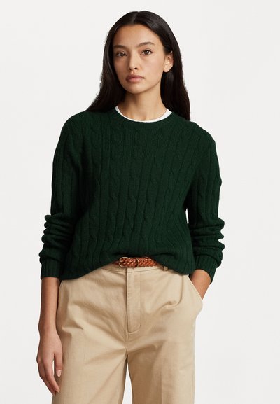 Pull en maille torsadée vert avec un col rond, doté de manches longues et de bord-côtes. Porté sur un pantalon beige avec une ceinture tressée.