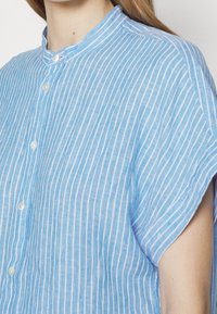 Blå randig kortärmad skjorta med mandarin-krage, med vita vertikala ränder och knäppning fram. Lättvikts tygstruktur.