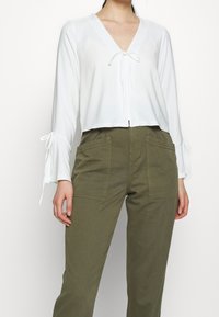 Blusa bianca, corta, con maniche lunghe e scollatura a V, con nastri ai polsini e chiusura anteriore a nodo. Abbinata a pantaloni verde oliva.