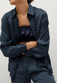 Frau, die die Arme verschränkt und ein dunkelblaues, florales Musterhemd sowie dazu passende Hose über einem gerafften schwarzen Bandeau-Top trägt.
