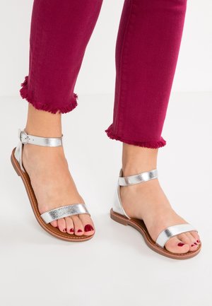 Srebrne metaliczne sandały z dwoma szerokimi paskami i klamrą przy kostce, zestawione z przyciętymi bordowymi dżinsami z postrzępionymi brzegami.