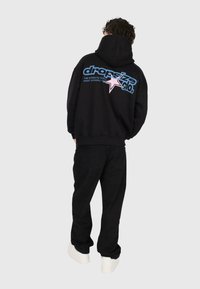 Person wearing black hoodie and pants, white shoes, hoodie back displays blue and pink text reading "dropsize clo." with a star graphic.