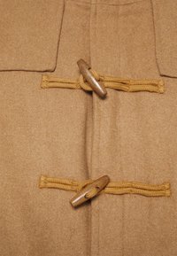 Beige Wollmantel mit Holz-toggle-Verschlüssen und geflochtenen gelben Bändern, glatte Textur und Stoff, der Filz ähnelt.