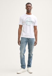 Hvid bomulds t-shirt med grafisk print, der har tekst og cirkulært design. Båret med lyseblå denim jeans og hvide sneakers. Enkel, afslappet stil.