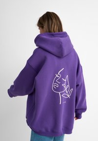 Felpa viola realizzata in tessuto morbido, caratterizzata da un grande design a contorno bianco di un volto e foglie sulla schiena. Vestibilità rilassata con cappuccio.