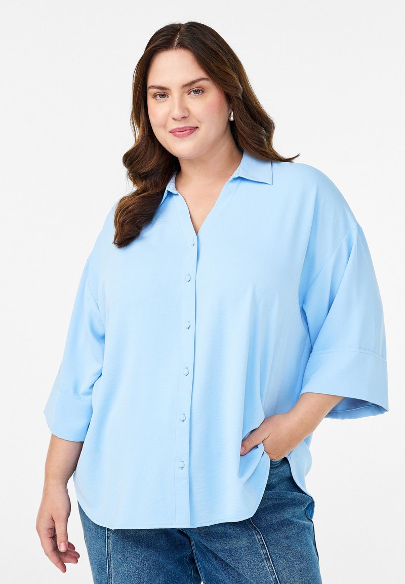 Woman with long brown hair wearing a loose light blue button-up shirt and blue jeans, posing with one hand in pocket on white background.