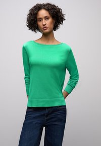 Femme aux cheveux bouclés portant un pull vert vif à manches longues et un jean bleu foncé, debout les mains derrière le dos.