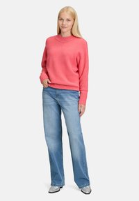 Vrouw met lang blond haar, gekleed in een roze sweater, lichtblauwe wijde jeans en zilveren puntige schoenen, staande met één hand in haar zak.