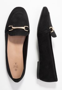 Mocassins en daim noir avec détail en métal doré, semelle plate et semelles intérieures beige portant les inscriptions "CALL IT SPRING" et "VEGAN." Vue de dessus de la chaussure gauche, vue de côté de la chaussure droite.