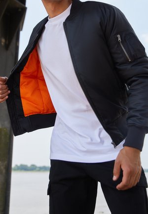 Person trägt eine schwarze Bomberjacke mit orangefarbenem Futter, weißem T-Shirt und schwarzer Hose und steht im Freien in der Nähe von Wasser.