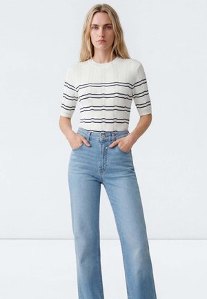 Frau mit langen blonden Haaren, die einen weißen kurzärmeligen Pullover mit schwarzen Streifen und hoch taillierte hellblaue Jeans trägt, Hände in den Taschen.