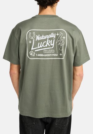 Camiseta de algodón verde con un diseño gráfico en la parte posterior que incluye el texto "Naturally Lucky Club" y ilustraciones de un herradura, una mano y dados.