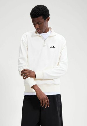 Uomo con una felpa bianche a mezza zip di ellesse che si sistema la manica, in piedi davanti a uno sfondo chiaro e uniforme.