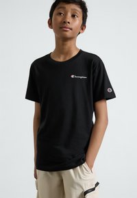 T-shirt en coton noir avec un col ras du cou, arborant un logo "Champion" rouge et blanc sur la poitrine gauche et la manche.