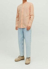 Camicia bottone color pesco chiaro con colletto alla cinese, abbinata a jeans di denim blu chiaro e scarpe beige, caratterizzata da un design semplice.